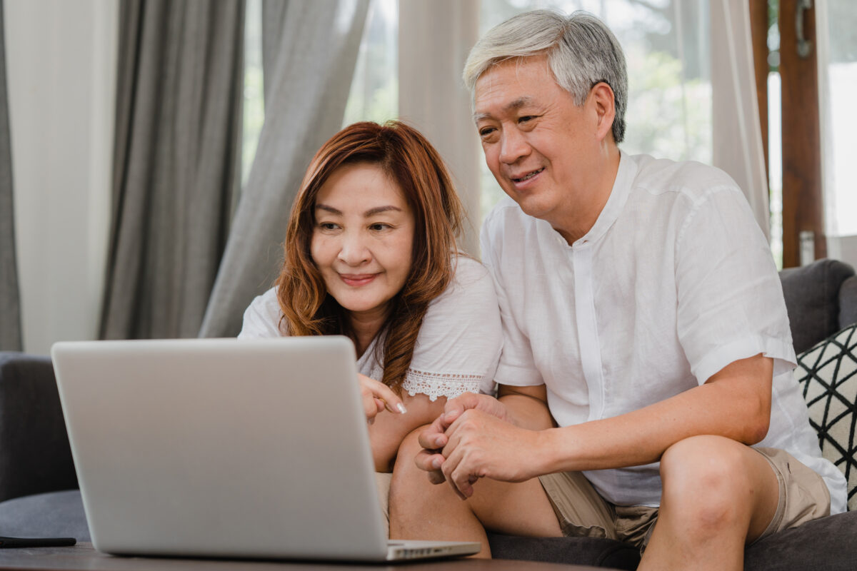 Asian senior couple using laptop at home. Asian Senior Chinese grandparents, surf the Internet to check social media while lying on sofa in living room at home concept.